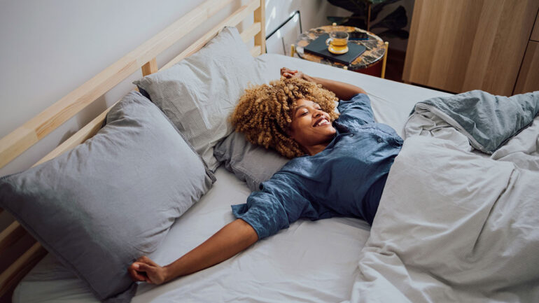A person with curly hair lies stretched out on a neatly made bed, smiling and relaxing under light bedding in a bright, modern bedroom.