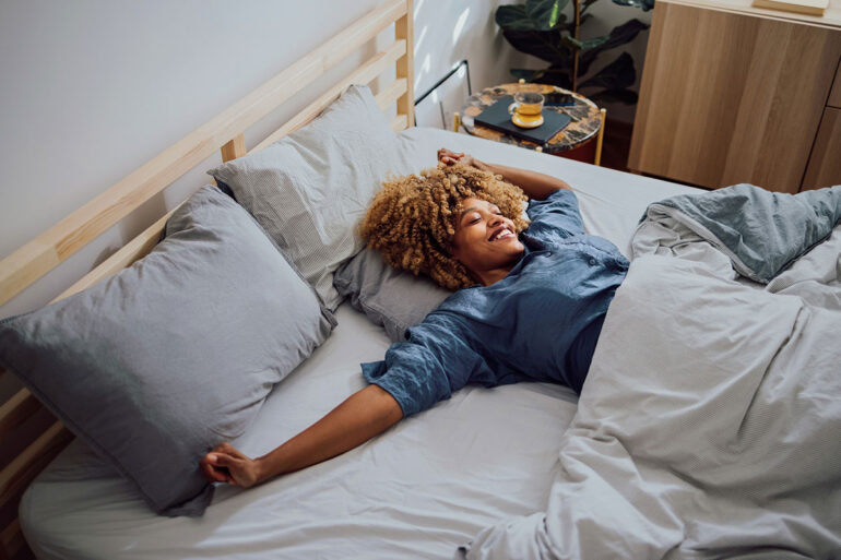 A person with curly hair lies stretched out on a neatly made bed, smiling and relaxing under light bedding in a bright, modern bedroom.