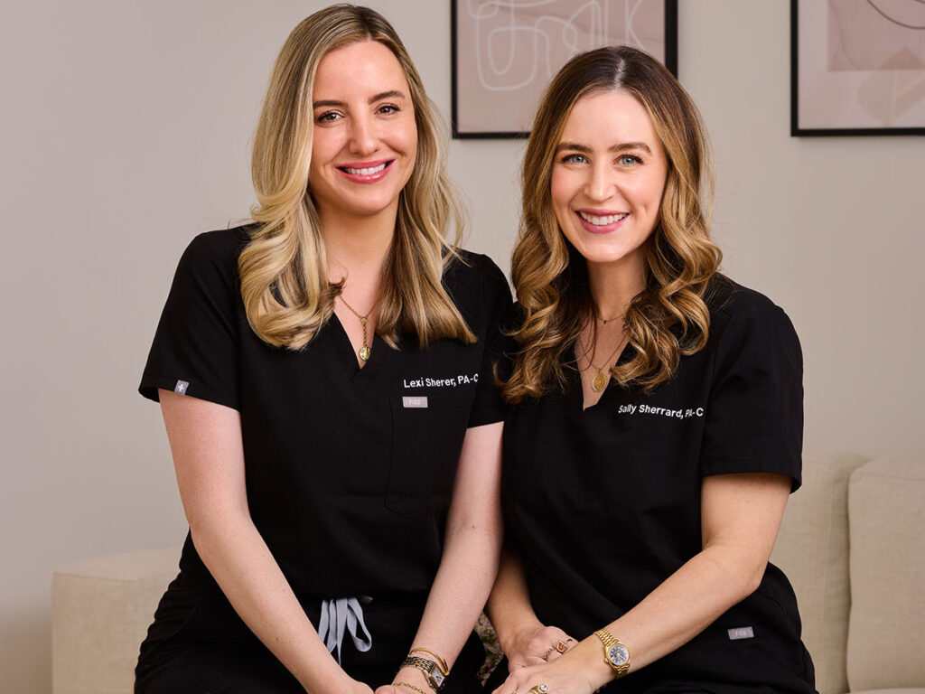 Two women wearing scrubs relax on a couch, smiling and enjoying each other's company.