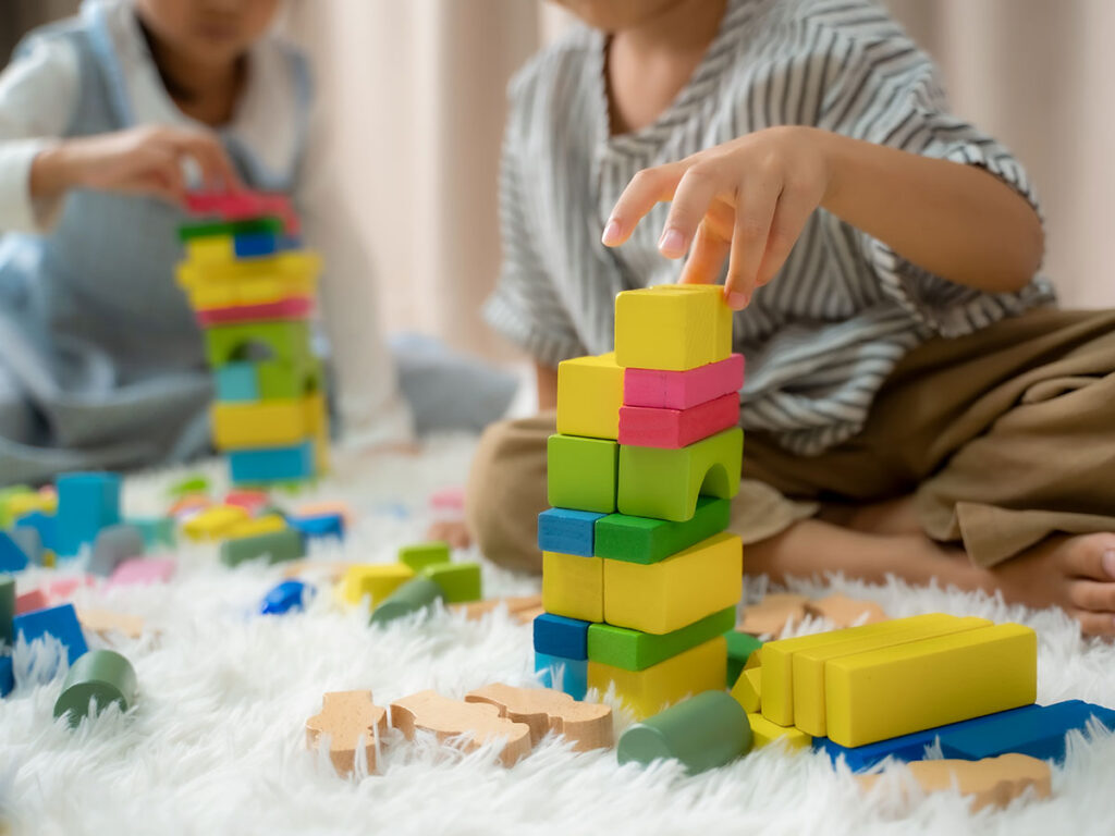 Two children play on a soft rug, building colorful towers with blocks. The scene conveys creativity and concentration in a cozy setting.