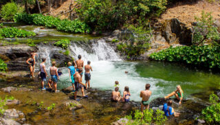 A group of kids in swimsuits enjoy a small waterfall and pool in a lush, green forest.