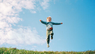 A child joyfully jumps on a grassy hill under a bright blue sky with scattered clouds, arms spread wide, conveying freedom and happiness.