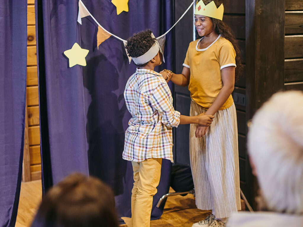 Two children in casual costumes perform on a stage with blue curtains and star decorations.