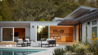 Modern house exterior with large windows, patio seating, and a pool. Warm indoor lighting creates a cozy atmosphere, surrounded by lush trees.