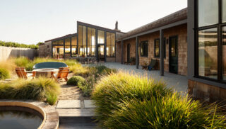 Modern house with large glass windows and wooden shingles, surrounded by lush grasses. A patio with chairs and a hot tub creates a serene, inviting setting.