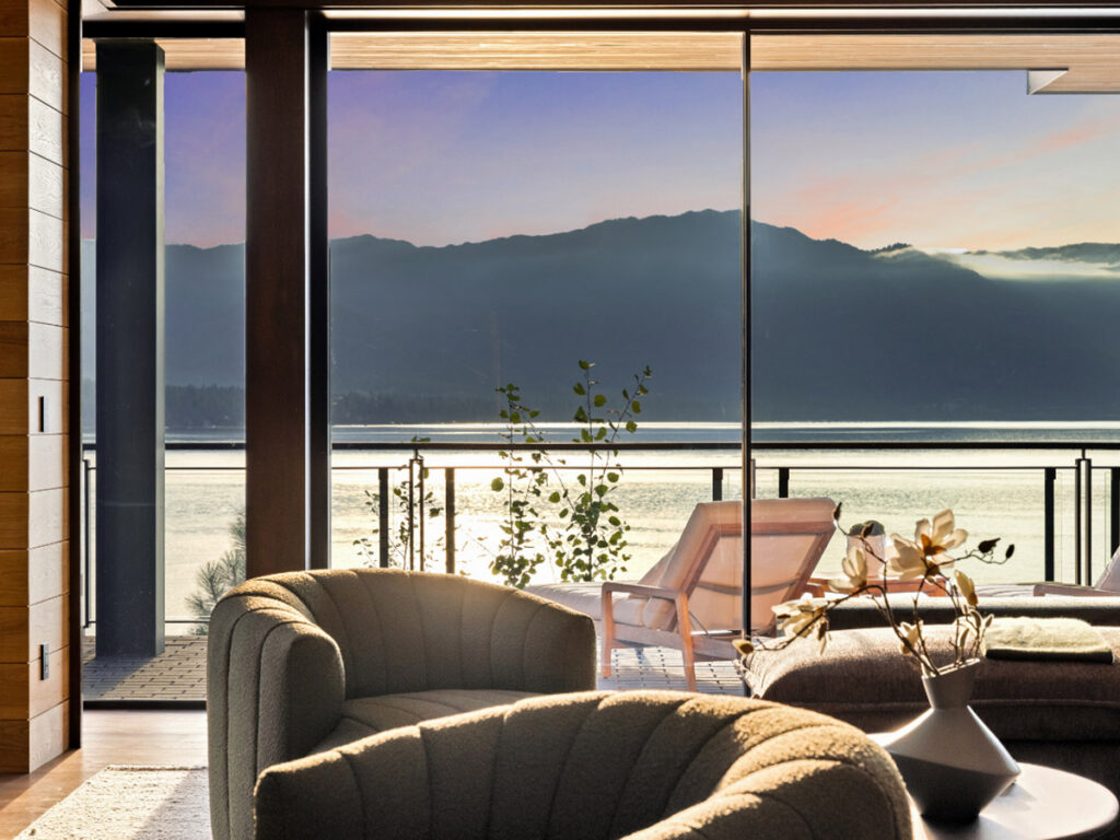 A contemporary living room with soft armchairs and a vase of flowers overlooks a tranquil lake and mountains at sunset through floor-to-ceiling windows.