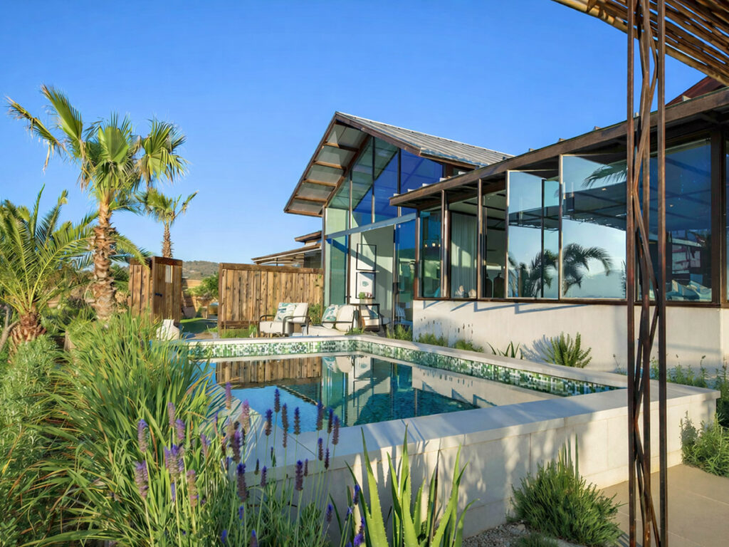 Modern house with large glass windows reflecting a clear blue sky, surrounded by lush greenery and a rectangular pool. Palm trees add a tropical vibe.