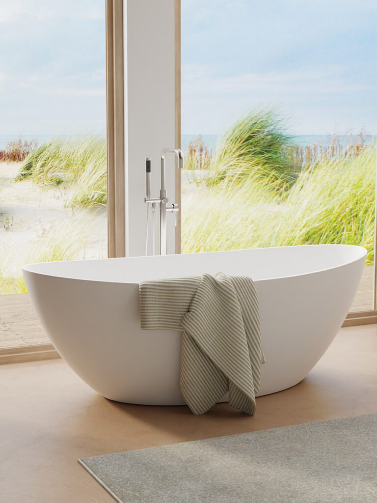 A white freestanding bathtub with a striped towel draped over the side sits in a room with large windows overlooking a serene beach scene with grassy dunes.