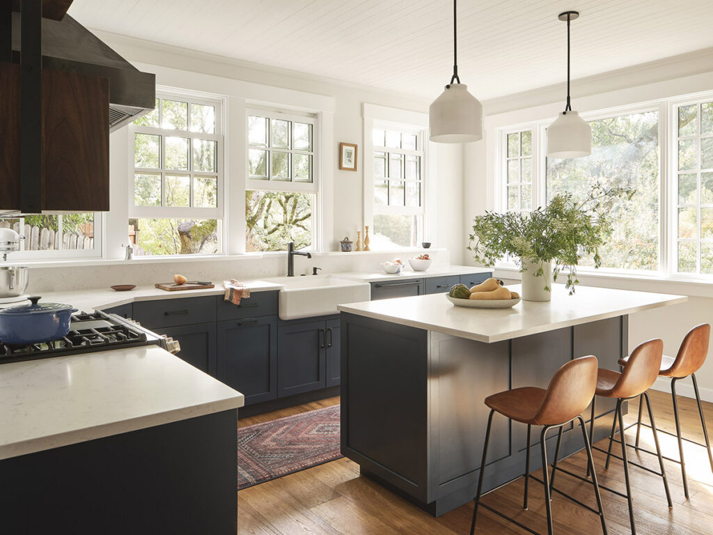 Bright, airy kitchen with large windows, navy cabinets, white countertops, pendant lamps, and a central island adorned with flowers and fruits.