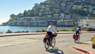 Cyclists ride along a coastal road under a clear blue sky. Across the water, a hillside is dotted with houses, creating a serene, picturesque scene.