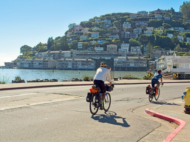 Cyclists ride along a coastal road under a clear blue sky. Across the water, a hillside is dotted with houses, creating a serene, picturesque scene.