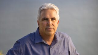 A man with light gray hair wears a blue button up shirt, standing in front of a gray background.