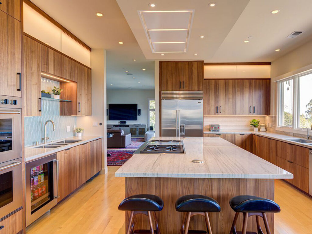 Modern kitchen with wooden cabinets, marble countertop island, and stainless steel appliances. Large windows and recessed lighting create a bright, airy atmosphere.