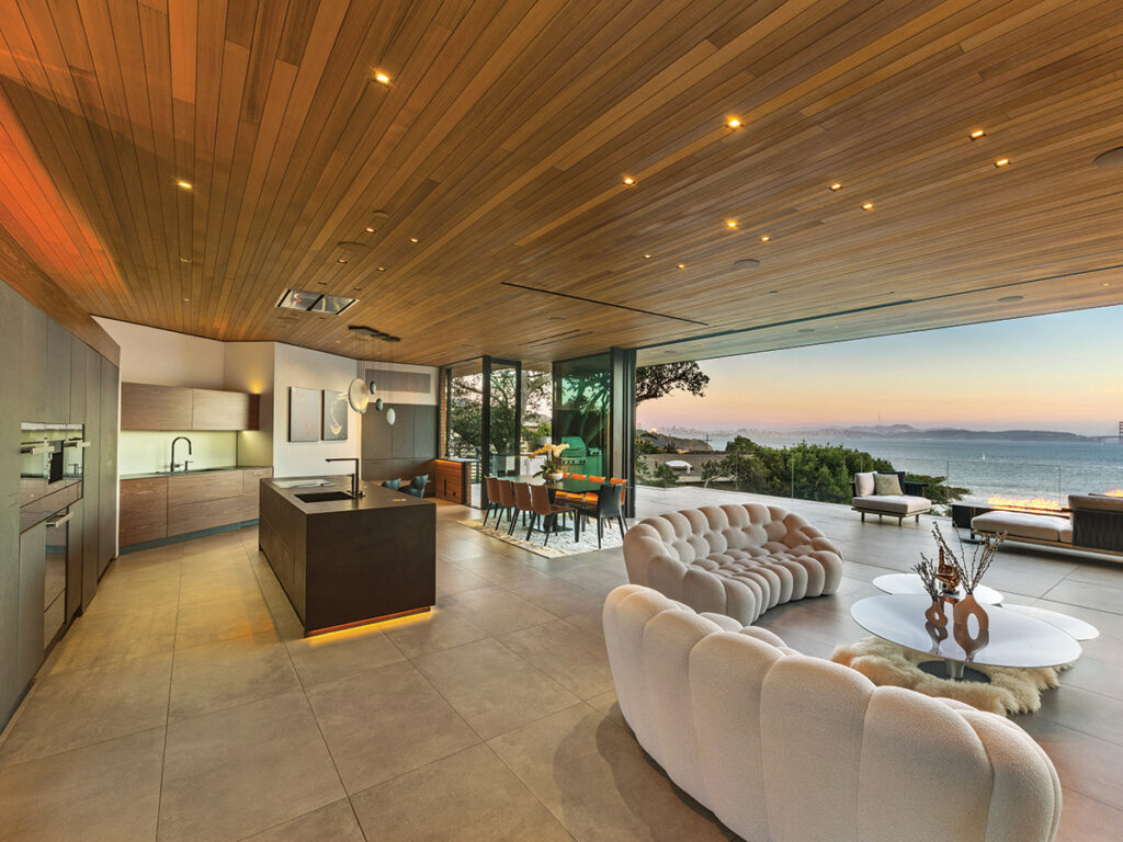 Luxurious open-plan living space with wooden ceiling, modern kitchen, stylish furniture, and floor-to-ceiling windows offering a serene ocean view at sunset.