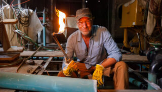 A person in work attire holds a lit blowtorch in a workshop, smiling confidently. The setting is industrial, with cables and machinery around.