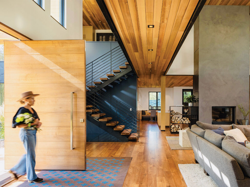 Modern home interior with wooden floors and ceiling, a woman carrying flowers enters. A sleek staircase and cozy living area with a fireplace are visible.