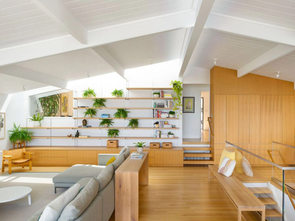 Modern living room with wooden floors, a light gray sectional sofa, and built-in shelving adorned with green plants and books. Bright and airy ambiance.