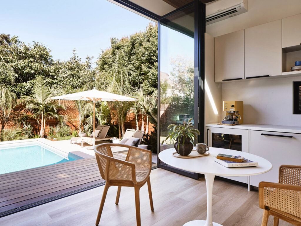 Modern room with a view of a sunny backyard featuring a pool, lounge chairs under an umbrella, and lush greenery. Cozy indoor seating and a coffee maker.