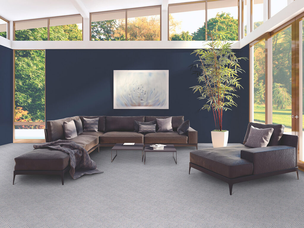 Modern living room with a large, dark sectional sofa, a central coffee table, and a tall potted plant. Large windows show greenery outside.