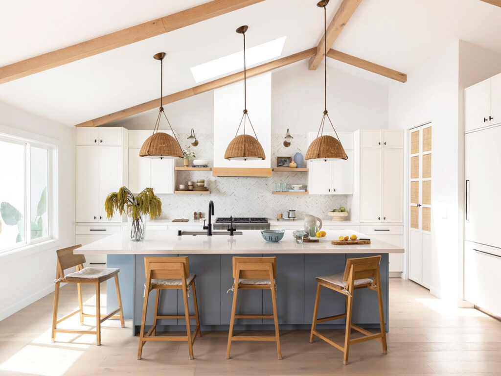 Bright, airy kitchen with vaulted ceiling and wooden beams. A large island with wooden chairs, woven pendant lights, and minimal decor create a cozy, modern feel.