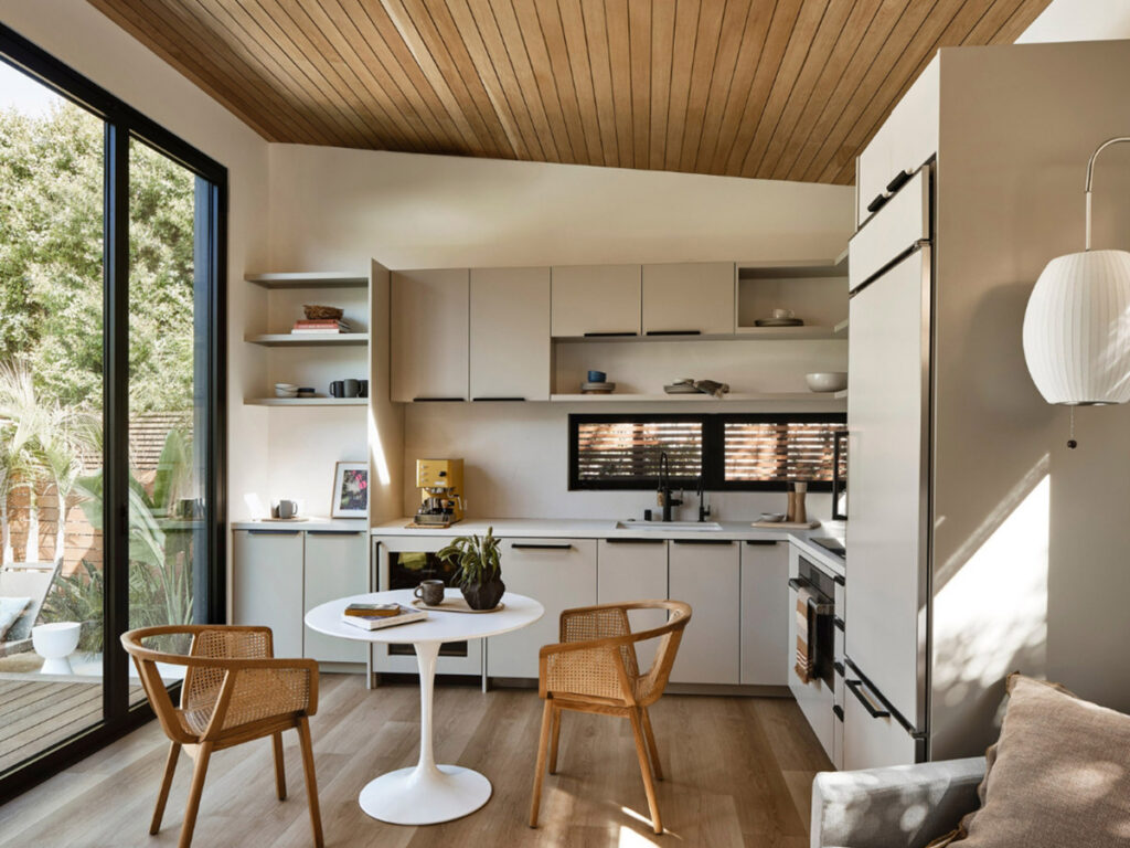 Stylish kitchen with sleek beige cabinets and a small round white table with two wicker chairs. Large window view of greenery, giving an airy feel.
