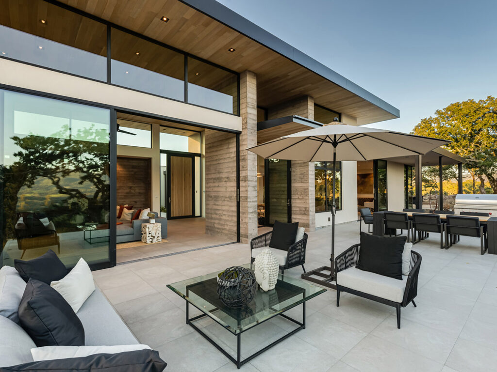 Modern patio with sleek furniture, large umbrella, and glass doors leading to a stylish interior. Sunlit and tranquil ambiance with nearby trees.