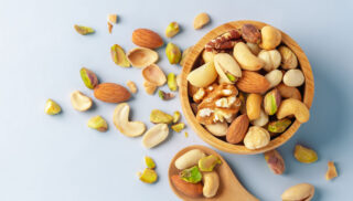 A wooden bowl filled with assorted nuts, including almonds, cashews, and walnuts, beside a wooden spoon with nuts scattered on a light blue background.