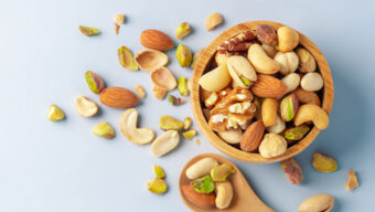 A wooden bowl filled with assorted nuts, including almonds, cashews, and walnuts, beside a wooden spoon with nuts scattered on a light blue background.