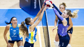 Two female volleyball players, one in blue and yellow, the other in purple, leap at the net, contesting the ball. Teammates watch, focused and intense.