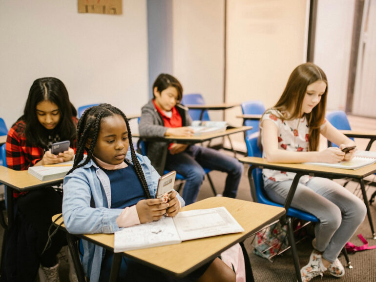 Four students sitting in a classroom, each focused on their phones.
