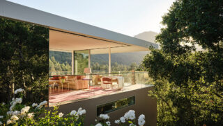 Modern glass-walled living room with pink decor overlooks lush forest, surrounded by white flowers and trees, exuding tranquility and elegance.