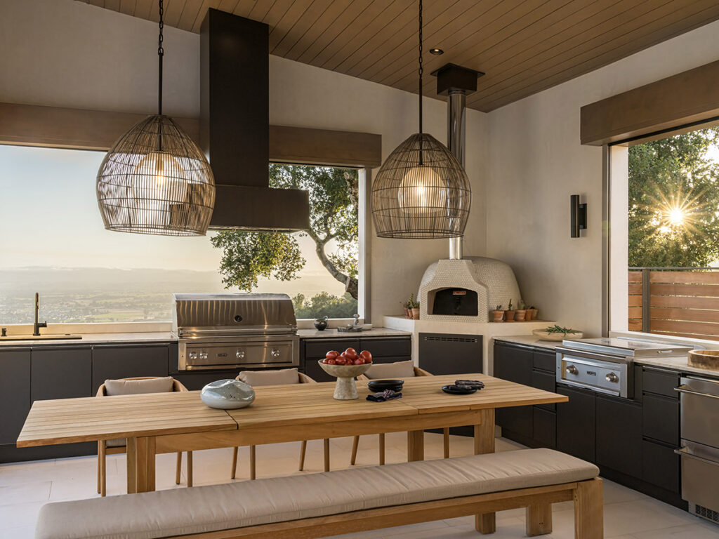 Modern outdoor kitchen with panoramic view. Features wood table, hanging wicker lights, stainless steel grill, and pizza oven.