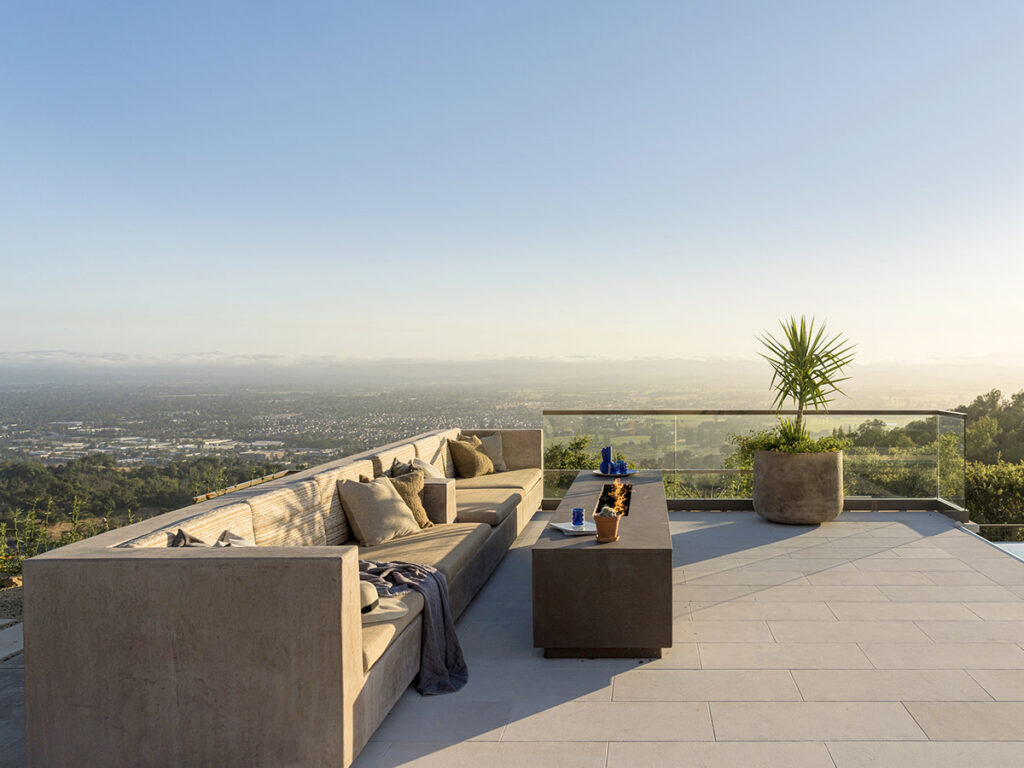Modern outdoor terrace with a beige L-shaped sofa adorned with cushions, a sleek gray coffee table, and a large potted plant. Scenic cityscape view under a clear blue sky.
