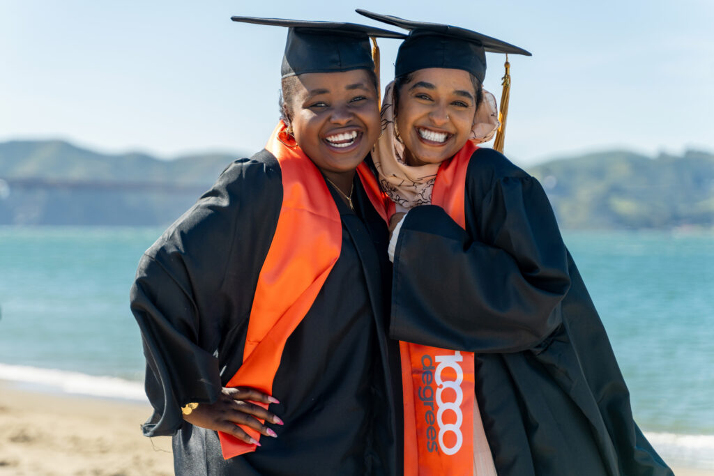 Two graduates wearing black caps and gowns with orange graduation sashes.