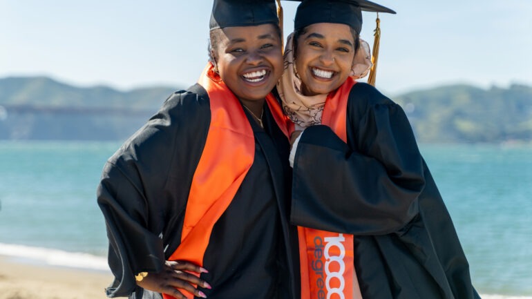 Two graduates wearing black caps and gowns with orange graduation sashes.