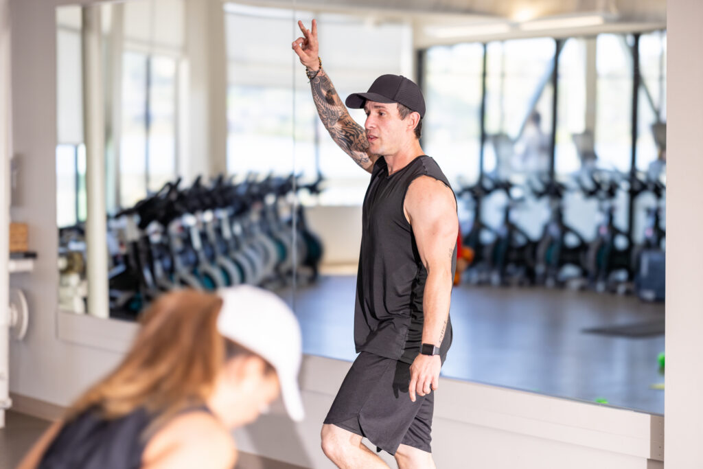 A male fitness coach, wearing a black hat, shirt and shorts, instructs a class.