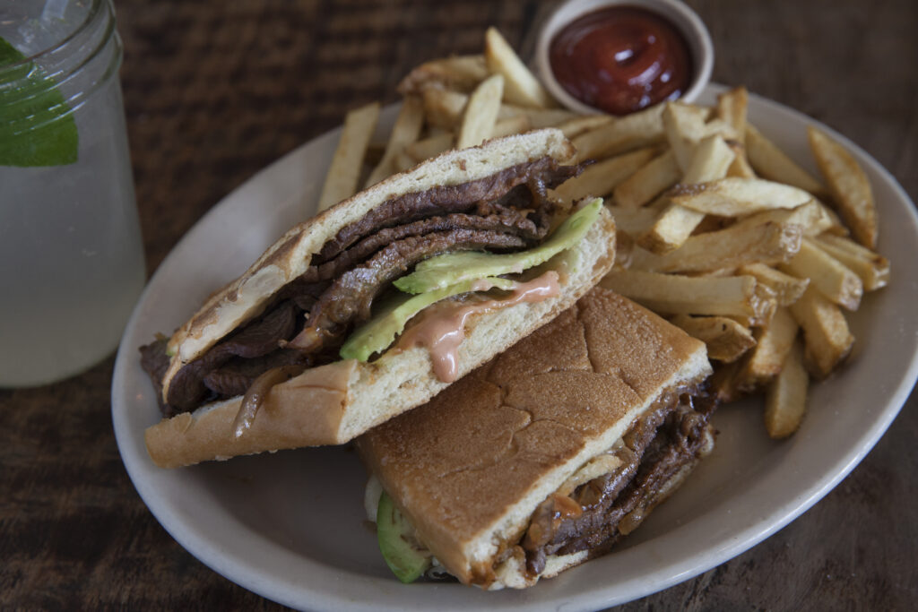 A steak sandwich, fries and ketchup served on a white plate. A glass jar filled with a clear liquid and mint leaf is off to the side.