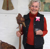 An older woman with short gray hair, wearing glasses, a red turtleneck, and a black vest, stands smiling while holding a drink in one hand and touching a wooden bird sculpture with the other. New Works are displayed on the wall behind her at ArtWorks Downtown.