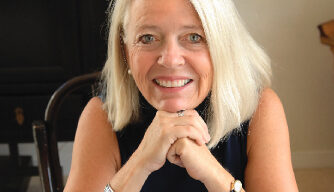 A woman with straight, shoulder-length blonde hair is smiling at the camera. Resting her chin on her clasped hands, she exudes a nonviolent aura. She wears a black sleeveless top, accented by a silver watch and thick silver bracelet. The indoor setting frames her serene vision perfectly.