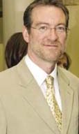 A man with glasses, a short beard, and short hair is wearing a light-colored suit, a white shirt, and a patterned tie. Standing indoors at McCall and Associates, another person is partially visible in the background.