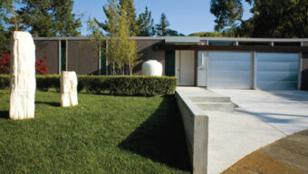 A Next-Century Modern single-story house with a sleek design has a gray facade, large windows, and a two-car garage. The well-manicured lawn features tall, abstract stone sculptures near the driveway. Trees and shrubs provide a natural backdrop to this contemporary home.