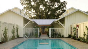 A serene swimming pool is centered between two modern barn-style buildings with white exteriors and pitched roofs. There's a large tree in the background, and the pool is enclosed by a fence, creating a peaceful setting that embodies Stewards of Change. The sky is clear.
