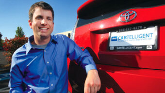 A man in a blue shirt poses next to the rear end of a red Toyota car, smiling. The car, an example of Alternative Buying, has a Cartelligent license plate frame, and the sun is shining brightly in the background.