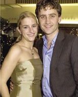 A smiling couple dressed formally stands close together, the woman in a tan strapless dress and the man in a dark suit with a blue shirt. They are indoors with soft lighting and decorative lights in the background, evoking an elegant Oscar night ambiance.