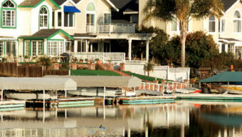 A serene lakeside neighborhood in Bel Marin Keys with large houses featuring gabled roofs and expansive windows, reflected in the still water. Various small boats and canoes are docked along the waterfront. Palm trees and manicured gardens add to the picturesque setting.