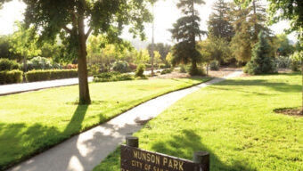 A sunlit park with neatly trimmed grass and a paved pathway curving through it. Tall trees provide shade, while a wooden sign in the foreground reads, "Munson Park, City of San Rafael, Closed Sunset to Sunrise." Just beyond lies the charming neighborhood of Terra Linda.