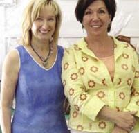 Two women, likely godmothers, stand and smile together. The woman on the left wears a blue sleeveless top, while the one on the right sports a yellow shirt with a floral pattern. They are in an indoor setting, with a sign that reads "Auction" partially visible in the background.