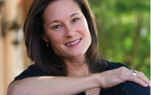 A woman with long dark hair, dressed in a black top, sits outdoors with a bright smile. She has one arm resting on her knee with a wooden structure behind her. The background is blurred, showcasing greenery. This moment captures the effortless grace reminiscent of Jennifer Azzi's iconic charm.