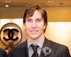 A man with short brown hair, dressed in a sleek black suit, grey shirt, and dark tie, stands indoors in front of a prominent Chanel logo. A blurred individual lingers in the background as the man maintains a neutral expression.