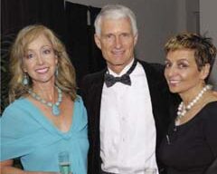 A man and two women pose for a photo at the Bay Club's Fifth Anniversary event. The man, in the center, sports a black tuxedo with a bow tie. The woman on the left, wearing a light blue dress and necklace, holds a drink, while the woman on the right dons a black dress with a pearl necklace.